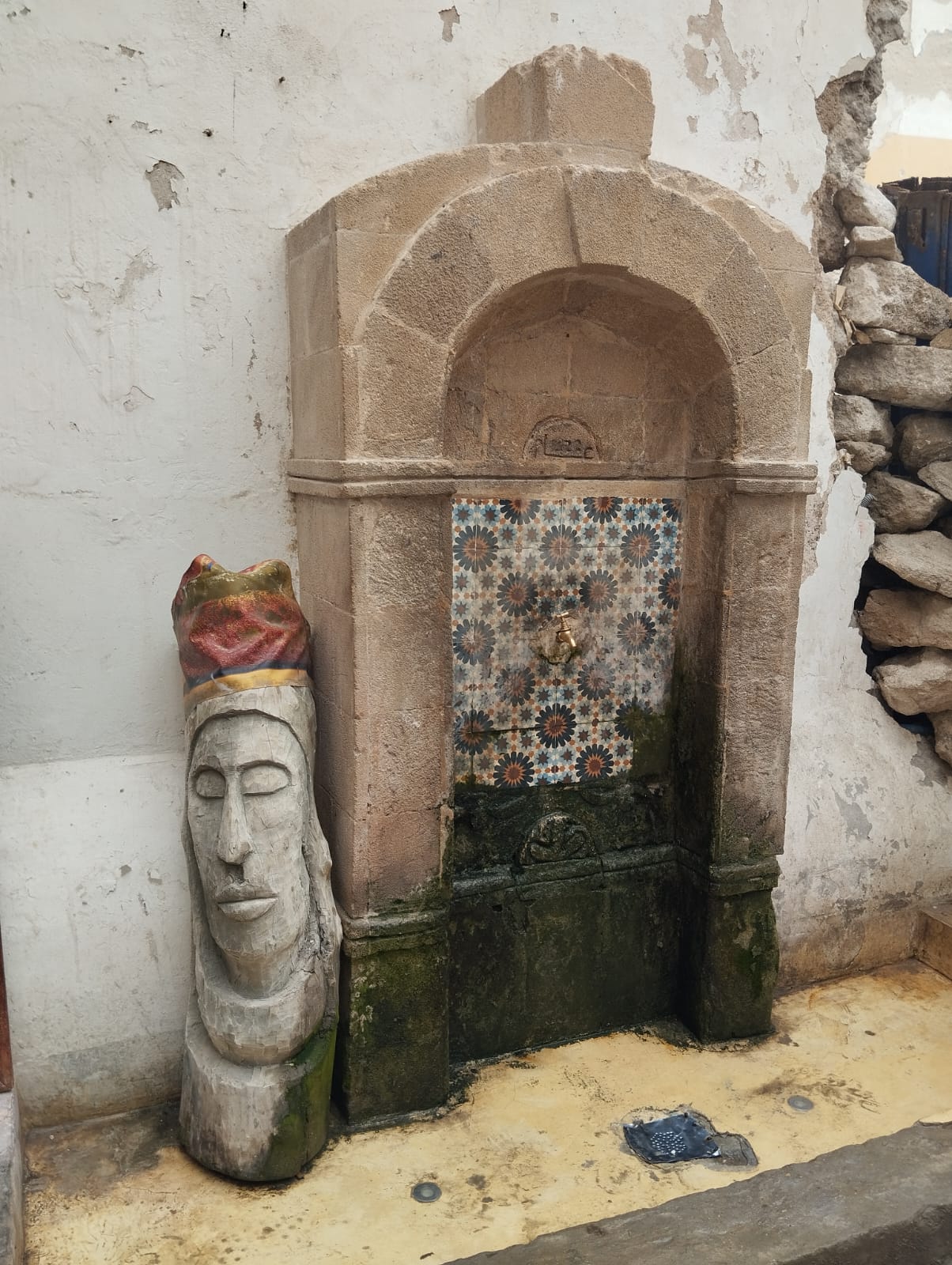 Ornate stone fountain with colorful tilework and a carved wooden head sculpture