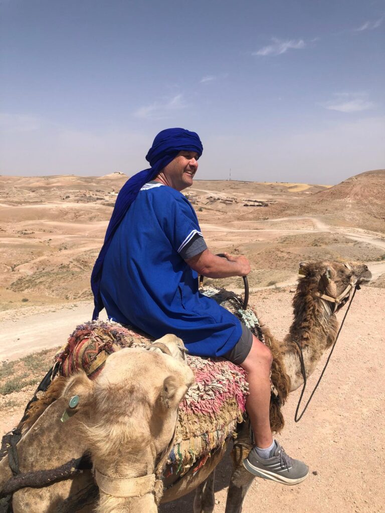 Man in blue turban and robe rides a camel in a desert landscape.