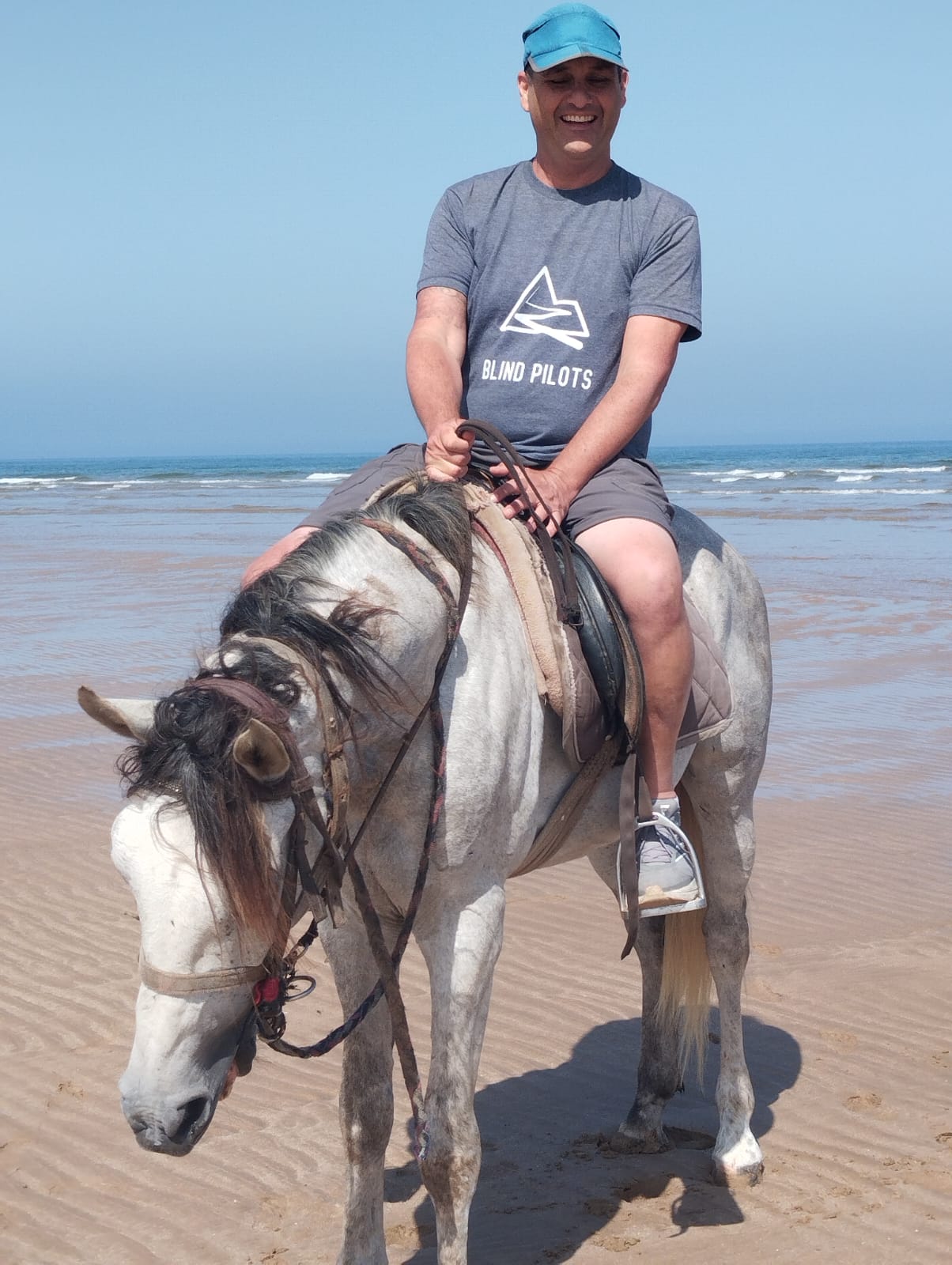 Man in "Blind Pilots" t-shirt rides a gray horse on a beach with ocean backdrop.