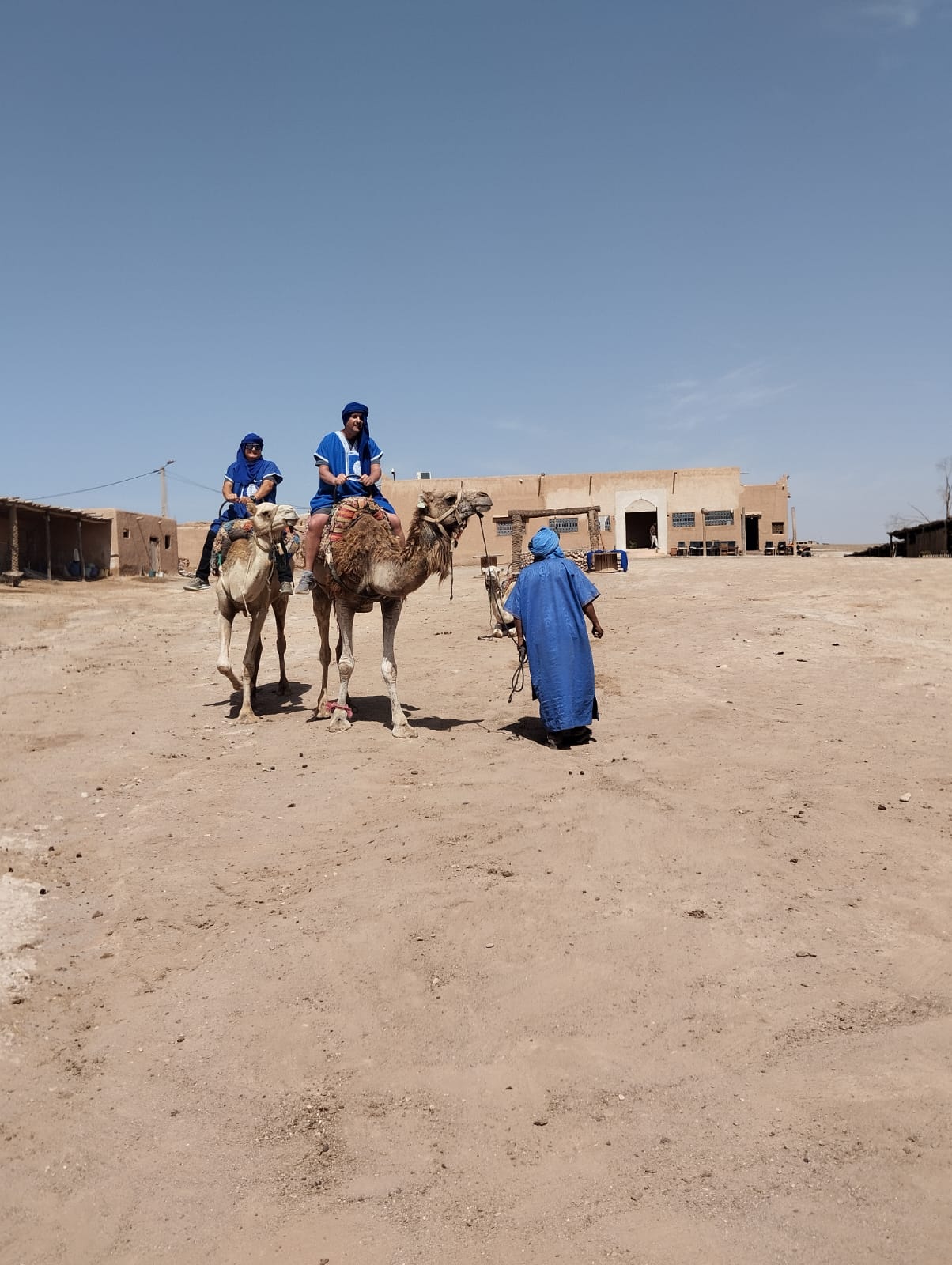 Two tourists ride camels in a desert village guided by a local.