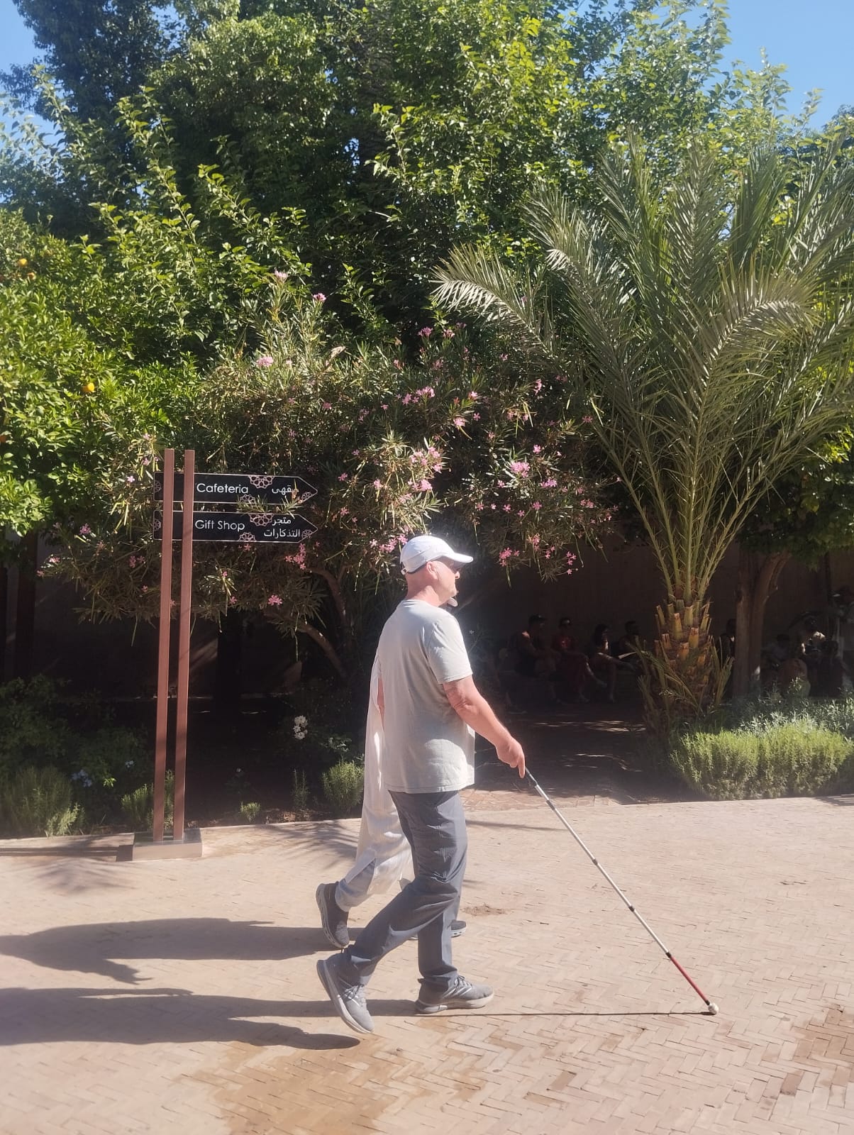 Man with white cane walks past signs for Cafeteria and Gift Shop in a garden.