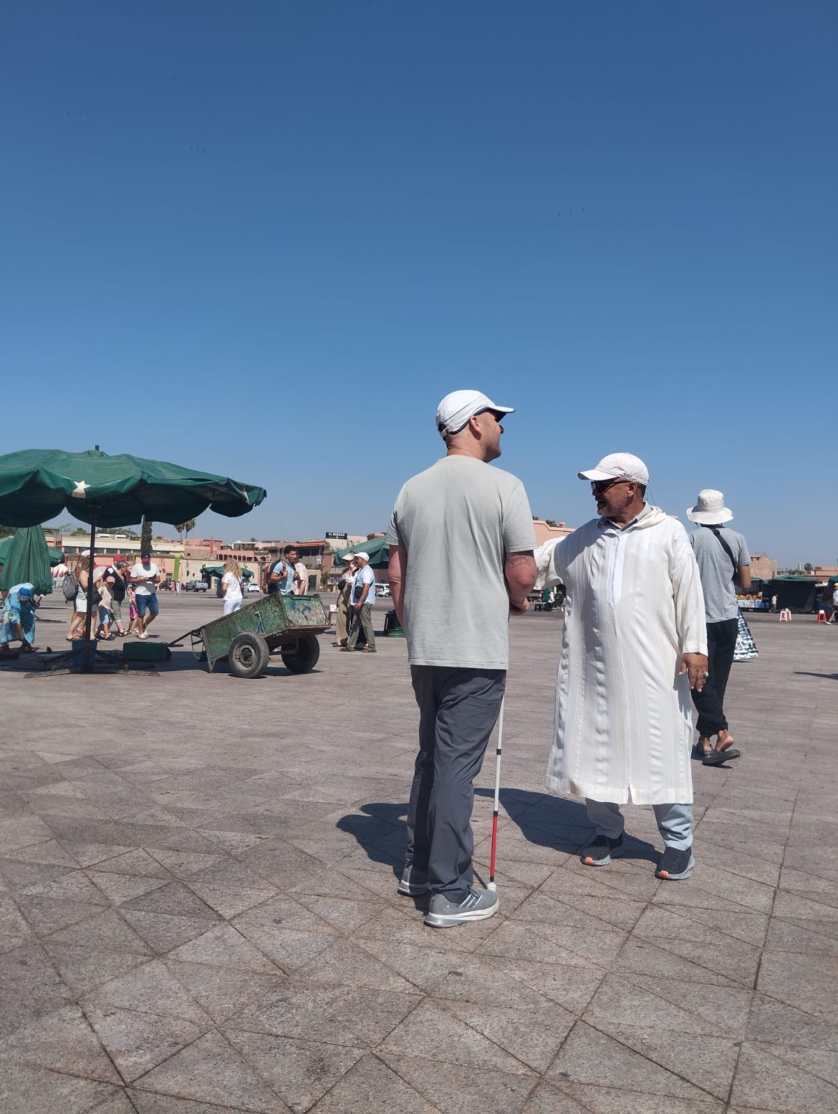Man with white cane talks to a man in a djellaba in a sunny Moroccan square.